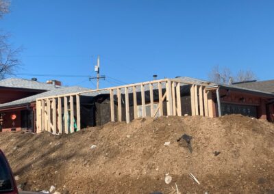 Home addition wall framing on a dirt-covered lot next to a brick ranch house