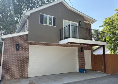 Two-story home addition with gray stucco upper level, brick lower level, and black balcony railing